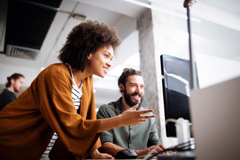Two people sitting behind computer screen smiling and talking